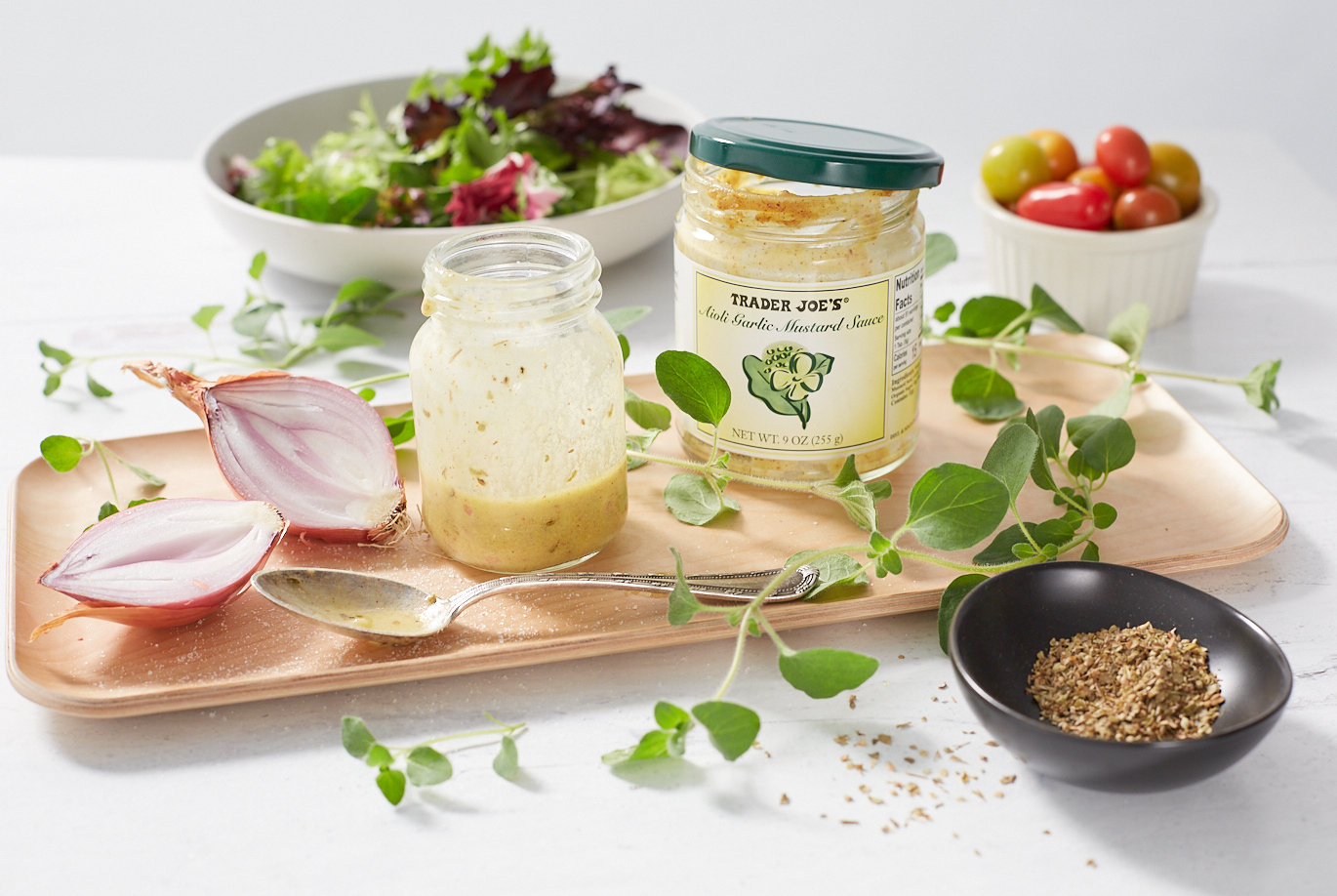 Trader Joe's Aioli Garlic Mustard Sauce used in dressing recipe with shallot and seasoning, on wood tray with fresh herbs, shallot and tomatoes in background