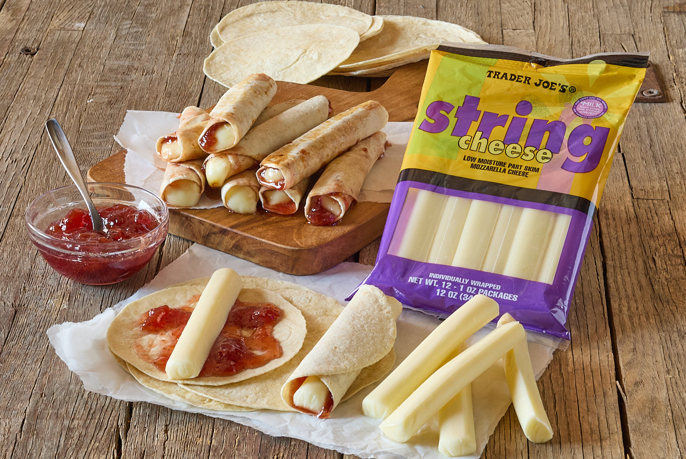 Trader Joe's String Cheese; shown in recipe for quick roll-up snack; mini tortillas on parchement, a wood board stacked with prepared roll-up snacks, and a small glass dish with strawberry preserves