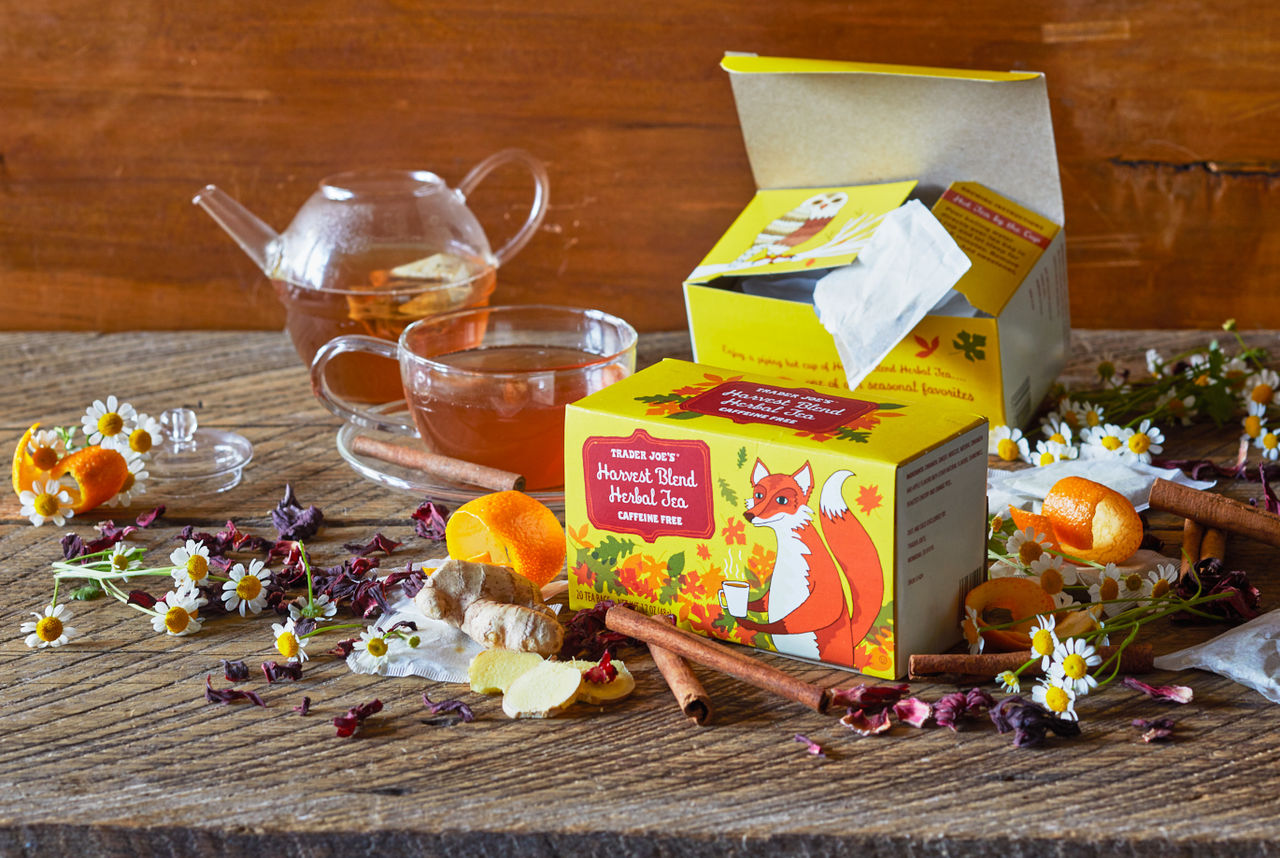 Trader Joe's Harvest Blend Herbal Tea box and packets surrounded by camomile flowers, orange peel, cinnamon sticks and dried hibiscus flowers; cup and teapot in background