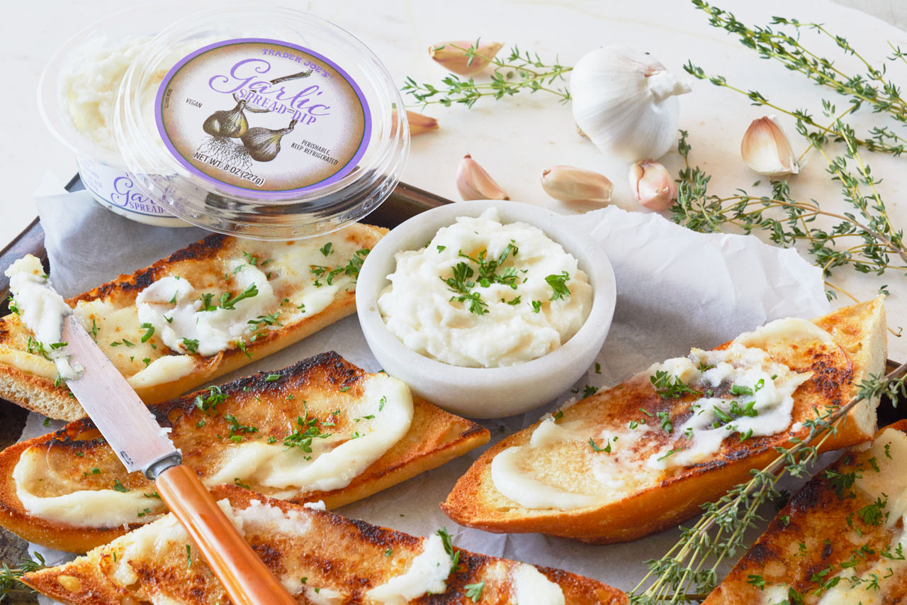 TJ's Garlic Spread Dip spread on toasted baguette slices, fresh garlic cloves and thyme surround pan