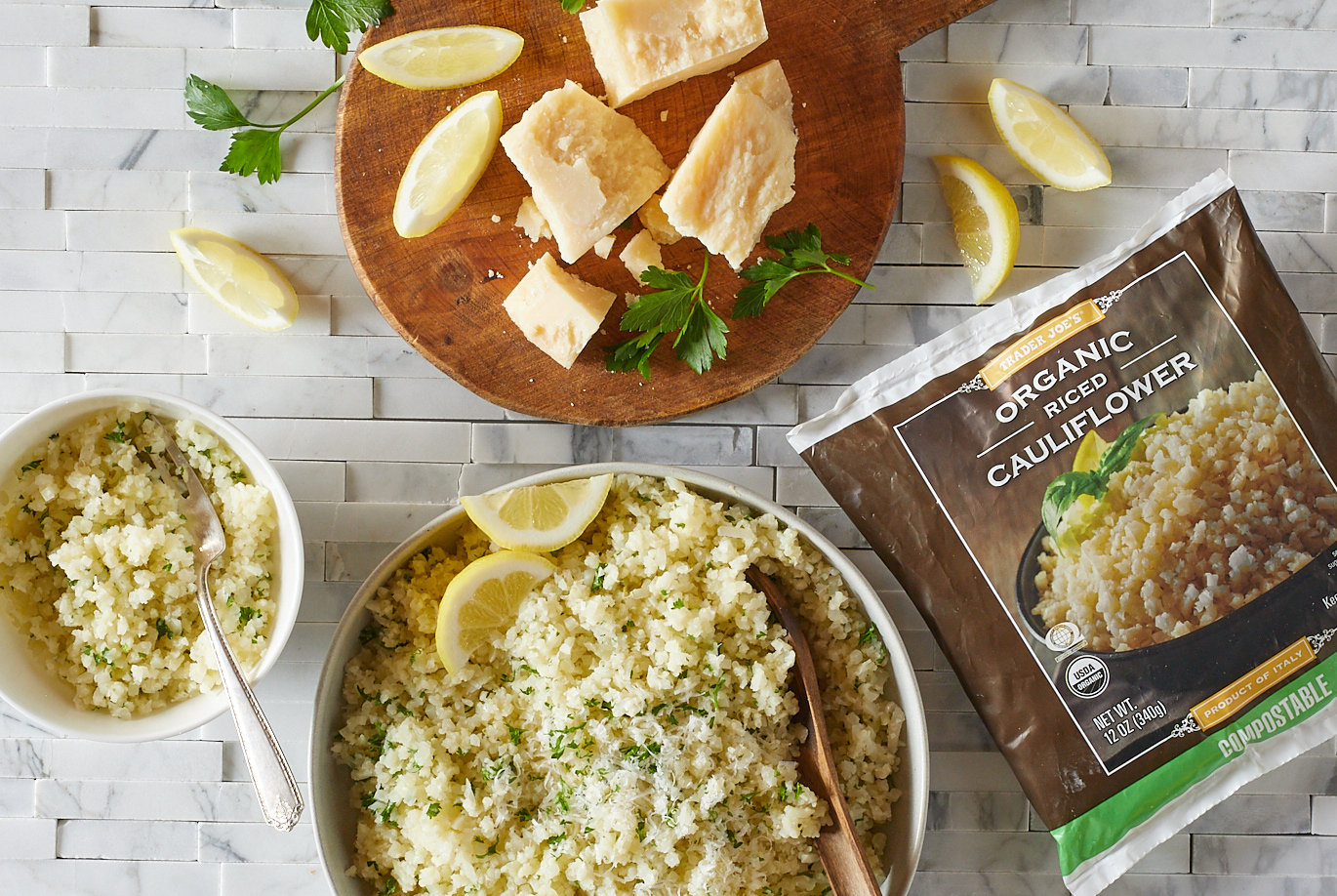 Trader Joe's Organic Riced Cauliflower used in recipe for Lemon Garlic Cauliflower Rice; prepared in a white bowl, with serving bowl on left; round wood board with chunks of Parmesan cheese, lemon wedges and parsley