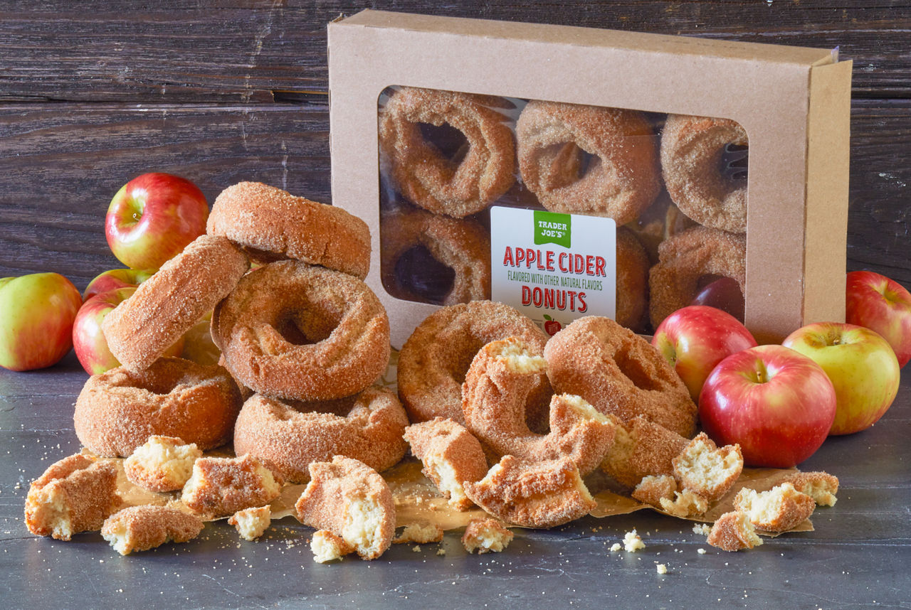 Trader Joe's Apple Cider Donuts; on dark background and surface, many donuts piled up ans some broken, apples surrounding