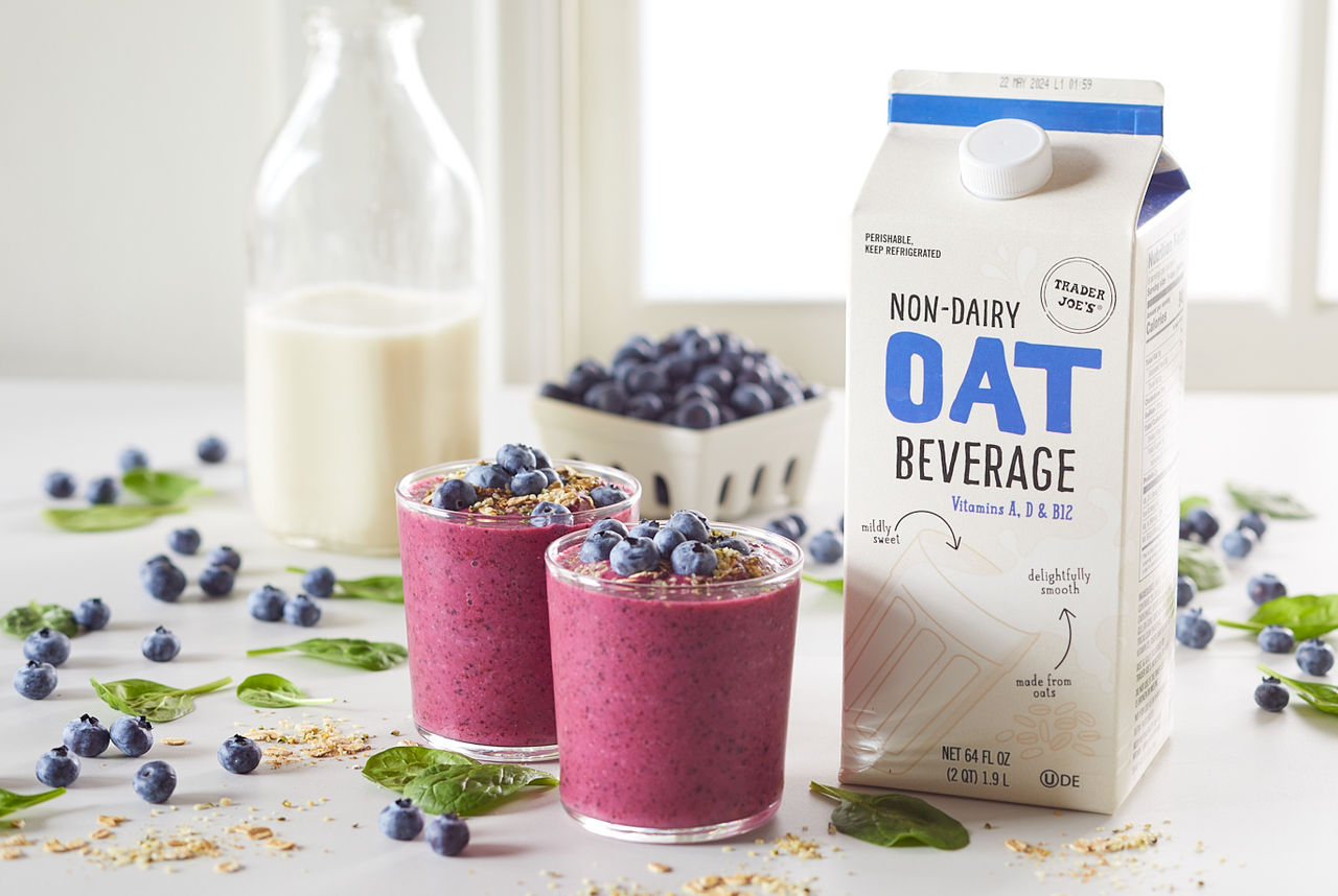 Trader Joe's Non-Dairy Oat Beverage, shown in recipe for Blueberry Protein Smoothie; two glasses with smoothie topped with fresh blueberries, hemp hearts and oats. Baby spinach, blueberries, hemp and oats scattered on surface