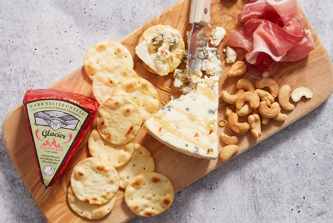 Carr Valley Wildfire Blue Cheese; on wood board, piece cut off and crumbled, surrounded byb crackers, cashews and prosciutto