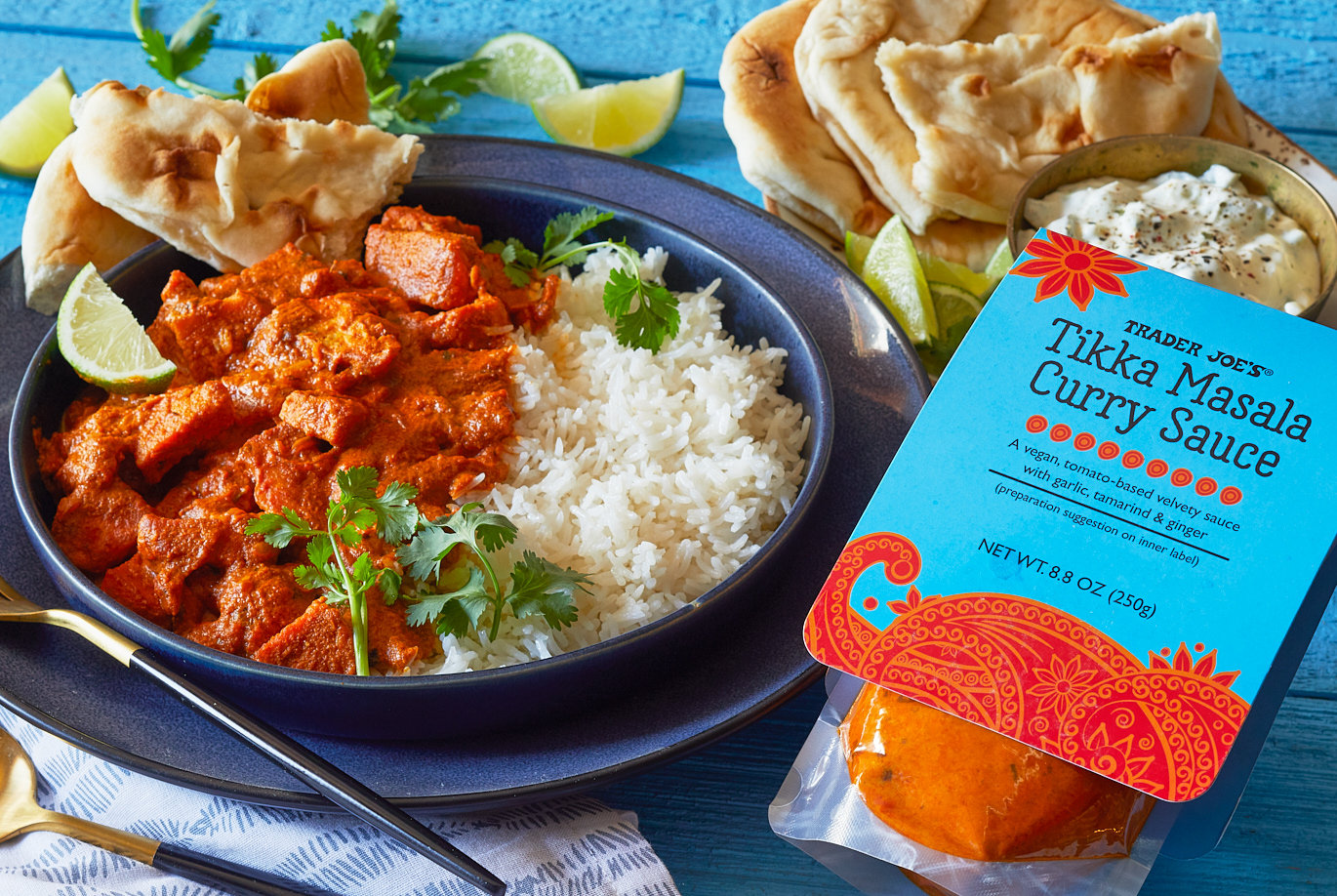 Trader Joe's Tikka Masala Curry Sauce; shown on blue surface and bowl; side by side with rice, garnished with parsley, lime and Naan bread