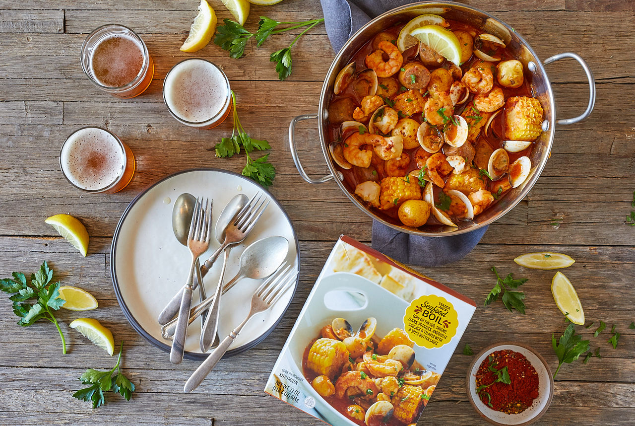 Trader Joe's Seafood Boil; prepared in stainless steel pot, a stack of plates and utensils on left, and several glasses of beer in background with lemon wedges and parsley