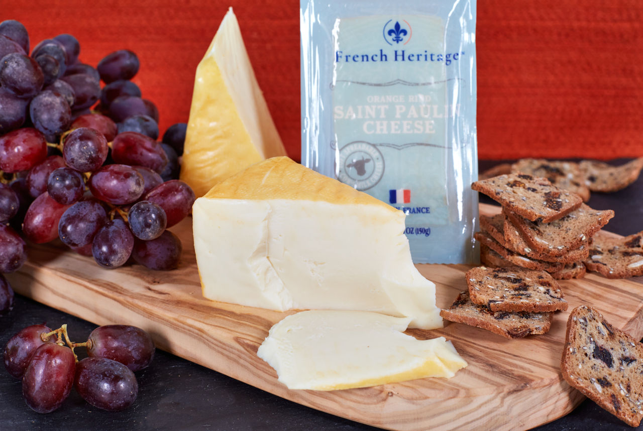 French Heritage Saint Paulin Cheese; on wood board, with red grapes and Fig & Olive Crisps; orange bacckground