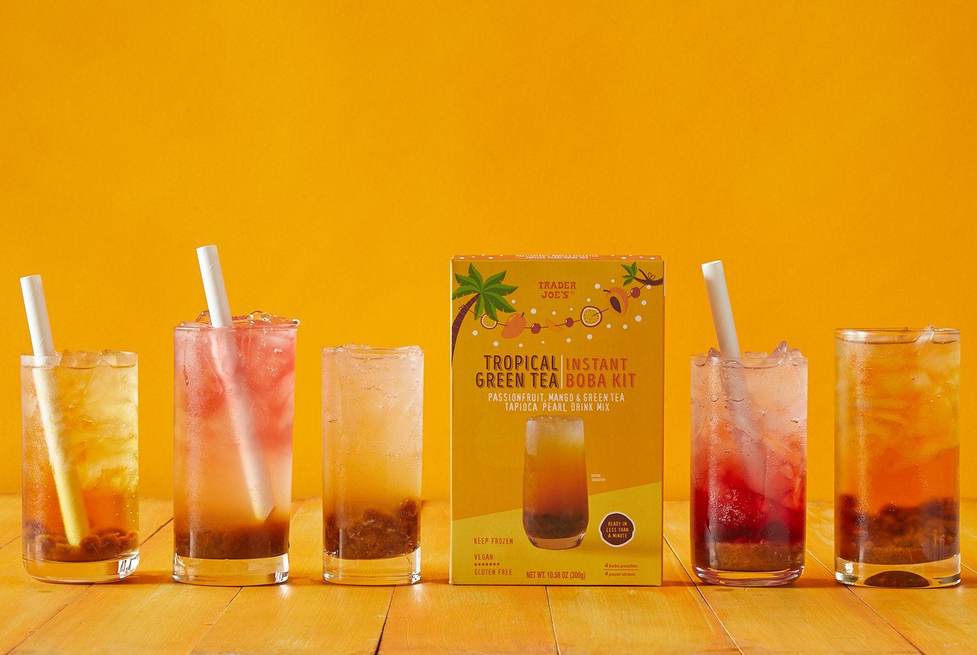 Trader Joe's Tropical Green Tea Instant Boba Kit; on bright orange backdrop, several tall glasses filled with ice, boba and a sparkling beverage, each a slightly different color