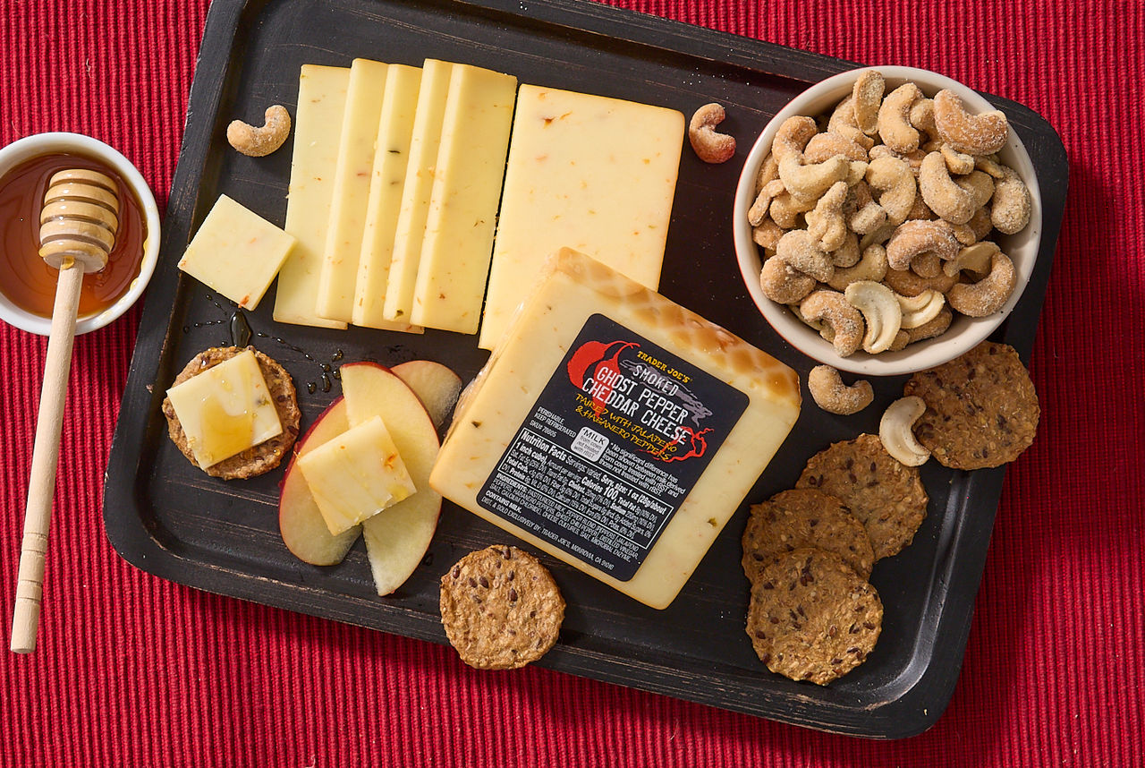 Trader Joe's Smoked Ghost Pepper Cheddar Cheese; shown sliced on a black tray, with seedy crackers, ranch cashews, apple slices and honey on red surface