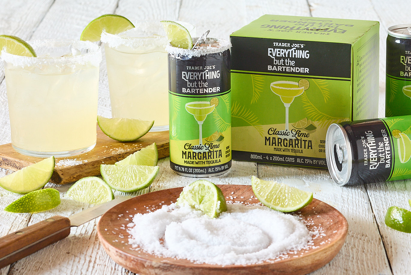 Trader Joe's Everything but the Bartender Classic Lime Margarita; many cans and a couple glasses; with salted rims and wedge of lime; more lime wedges on white wood surface along with wood plate of coarse salt